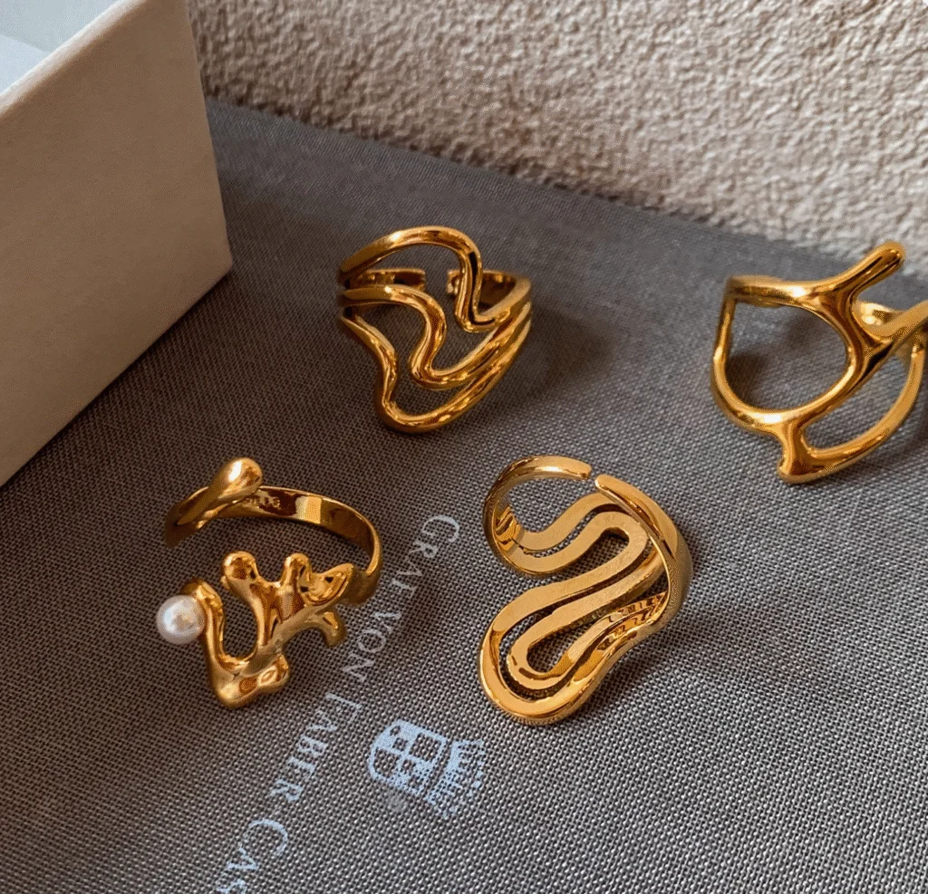 Four gold brass open rings with abstract and organic shapes, including one with a small pearl, displayed on fabric.